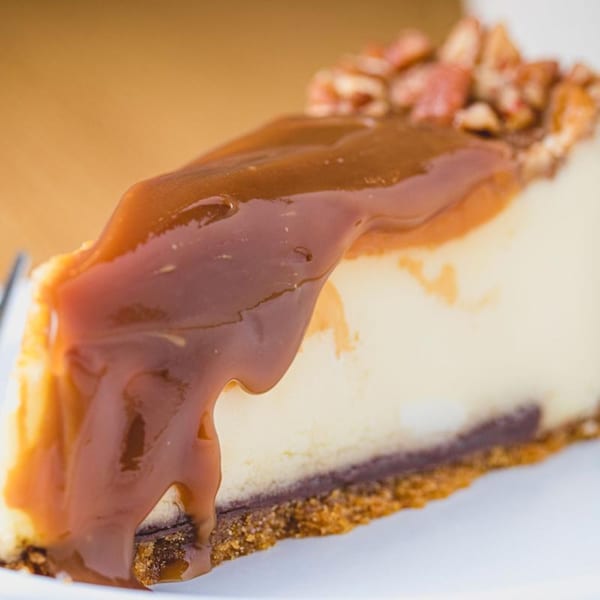 A slice of cheesecake with caramel drizzle and chopped pecans on a white plate.